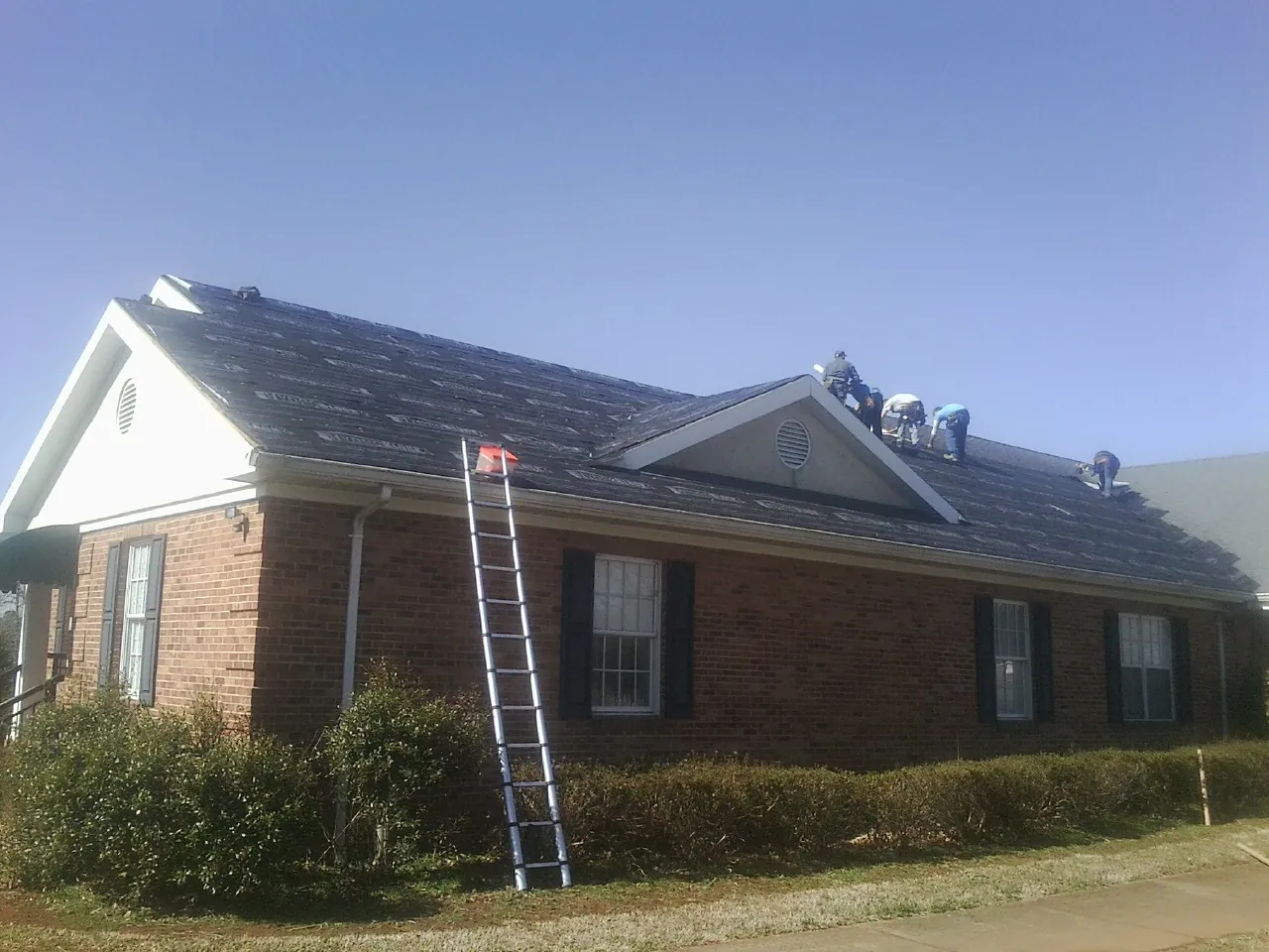 Crew tearing shingles off a brick ranch roof with a ladder set — Metal Roof Repair work by our crew in Olin