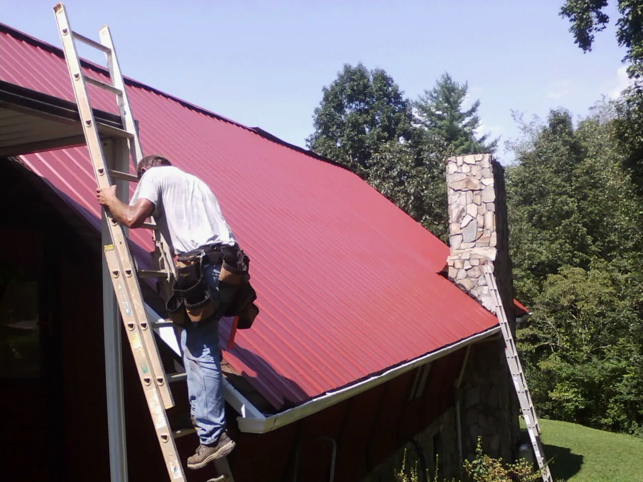 Roofer on a ladder beside a red metal roof and stone chimney — Soffit & Fascia Repair by Roofing Brothers in Olin
